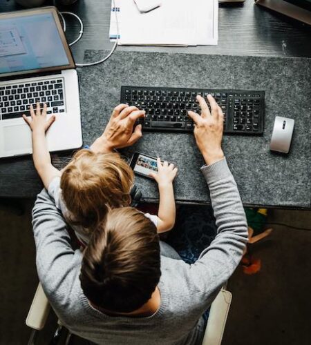 a man holds his son in his lap. they're both typing on different laptops